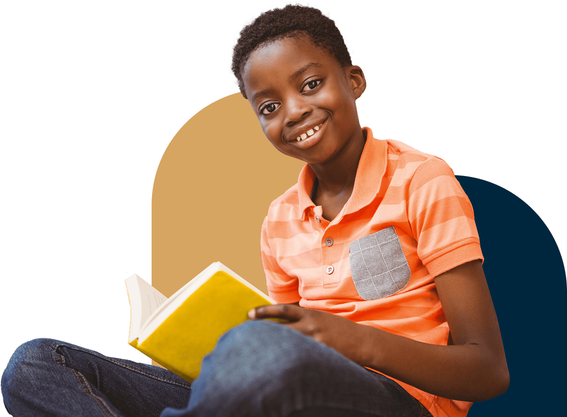 A smiling boy sits crossed-legged, holding a book and looking into the camera.