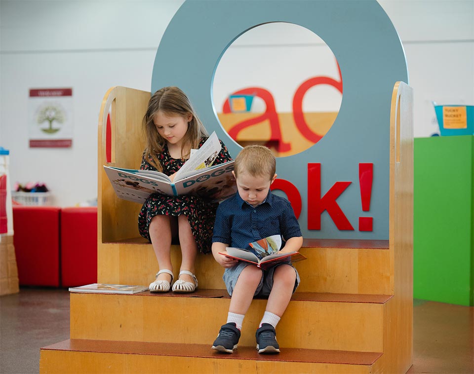 Two kids quietly focused on their books on fun wooden steps in the children's section.