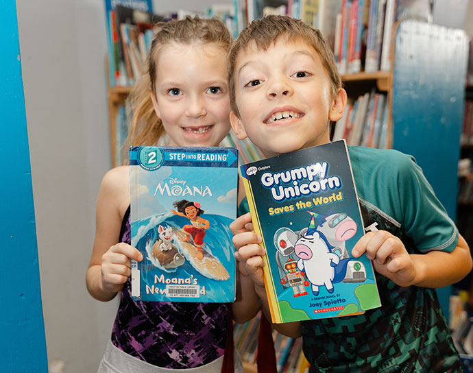 Two children smiling playfully and holding books.
